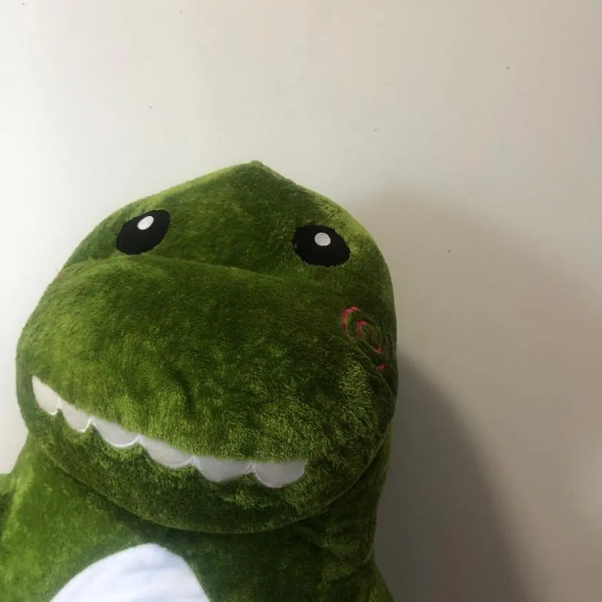 Dinosaur PFP #11: A fuzzy green dinosaur plush with round black eyes and a white toothy grin leans in close beside a plain white wall.