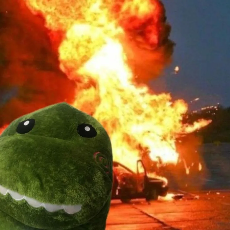 Dinosaur PFP #9: A green dinosaur plush with a white toothy grin stares into the camera while a car burns in bright orange flames behind it.